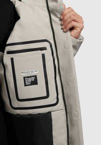 Interiorul unui sacou gri deschis, cu un buzunar cu fermoar și o etichetă rectangulară neagră. Designul include modele conturate în negru.