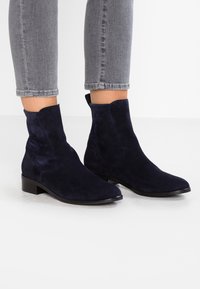 Bottines en daim bleu marine avec un talon carré bas, présentant une texture lisse, un bout arrondi et des panneaux latéraux élastiques.