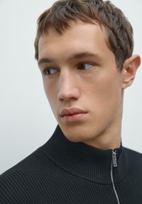 Giovane uomo con capelli corti castani che indossa un maglione nero a coste con zip, che guarda di lato su uno sfondo chiaro e semplice.