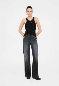 Zwarte tanktop met versieringen, gecombineerd met hoog-zittende, wijde, donkergrijze jeans. Model staat met handen op de heupen.