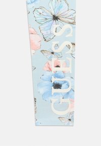 Μπλε ύφασμα με λουλουδένια σχέδια σε ροζ και μπλε, με πεταλούδες. Λογότυπο «GUESS» σε μεγάλο, κρεμ χρώμα με ελαφριά λάμψη.