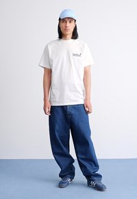 White cotton t-shirt with printed logo, blue cap, and loose-fitting blue denim jeans. Gray sneakers with white stripes. Simple, casual outfit.