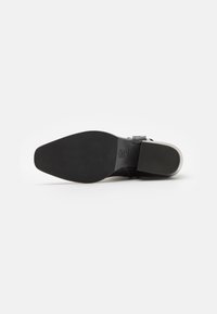 Chaussure en cuir noir avec une surface lisse, un bout pointu et une semelle en caoutchouc noir robuste. Détails incluent une sangle au milieu du pied.