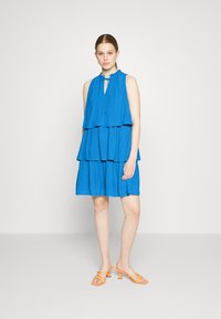 Vestido azul sem mangas com um design em camadas, apresentando um decote franzido e um padrão de pontos subtil. Combinado com sandálias de salto laranja.