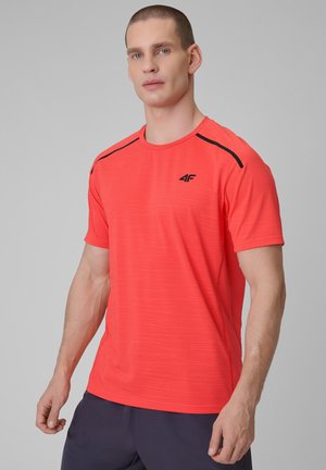 Mężczyzna w jaskrawoczerwonej sportowej koszulce z czarnymi paskami na ramionach i czarnym logo, w połączeniu z ciemnymi szortami, stoi na jednolitym tle.