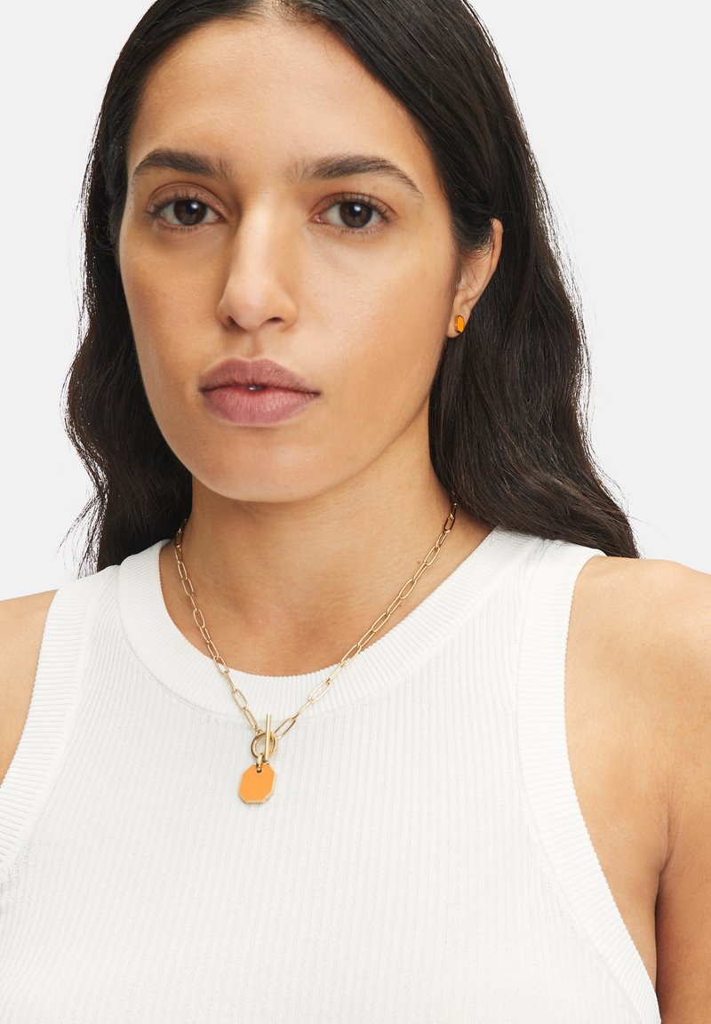 Gouden ketting met een oranje achtzijdige hanger, gedragen over een wit geribbeld tanktop, aangevuld met kleine oranje studoorbellen.