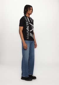 T-shirt negru din bumbac cu text și logo alb îndrăzneț, asortat cu blugi lejeri de denim bleu deschis și teniși negri.