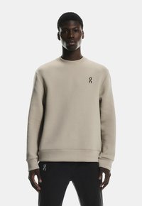 Hanorac bej deschis, cu guler rotund, manșete și tiv cu model texturat. Prezintă un mic logo negru pe piept. Textura materialului este netedă.