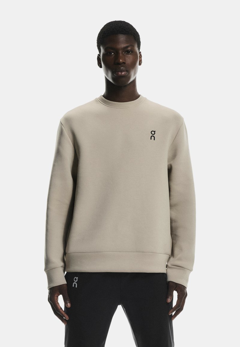 Hanorac bej deschis, cu guler rotund, manșete și tiv cu model texturat. Prezintă un mic logo negru pe piept. Textura materialului este netedă.