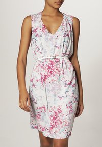 Frau in einem ärmellosen, knielangen Kleid mit weißem Grund und einem pixelierten Blumenmuster in Rosa, Lila und Blau, tailliert mit einem weißen Gürtel.