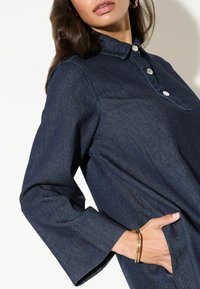 Tume teksasärg krae, nööpte ja pikkade varrukatega. Sellel on tasku ja sile tekstuur, mida rõhutavad õmbluste detailid.