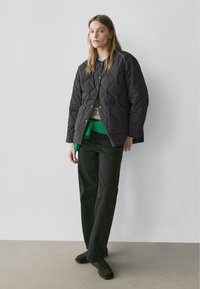 Ung kvinde står op ad en ensfarvet væg iført en sort quiltet jakke, grøn sweater bundet om taljen, mørke bukser og sorte sko.