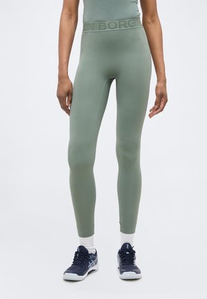 Egy magas személy, aki zsályazöld, magas derekú leggingset, fehér bokazoknit és tengerészkék sportcipőt visel, egy egyszerű háttér előtt áll.