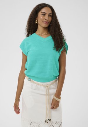 Hosszú, göndör hajú nő türkiz rövid ujjú kötött felsőben, fehér szoknyában díszített övvel, arany fülbevalóval és karkötővel szürke háttér előtt.