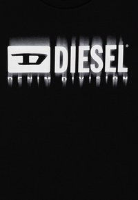 Juodas medvilnės marškinėliai, ant kurių dideliu balta grafika pavaizduota žodis "DIESEL" su stilizuotu logotipu ir "DENIM DIVISION" žemiau, parašyta ryškiu šriftu.