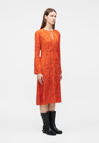 Γυναίκα όρθια, φοράει πορτοκαλί φόρεμα μήκους μέχρι τη μέση της γάμπας με μακριά μανίκια και σχέδια παϊνσλεϊ, καθώς και μαύρες μπότες μέχρι τη μέση της γάμπας, μπροστά σε λευκό, απλό φόντο.