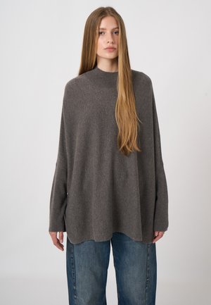 Woman with long straight hair wearing a loose gray sweater and blue wide-leg jeans standing against a plain light background.