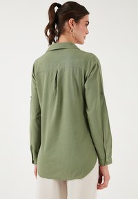 Svijetlozelena košulja dugih rukava s leđnim jaramom, zakrivljenim rubom i detaljem za podizanje rukava; mekana, teksturirana tkanina.