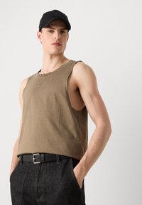 Brun uten ermer bomullstopp med rund halsåpning, med en liten logo; sammen med mørke denimbukser og et svart belte.