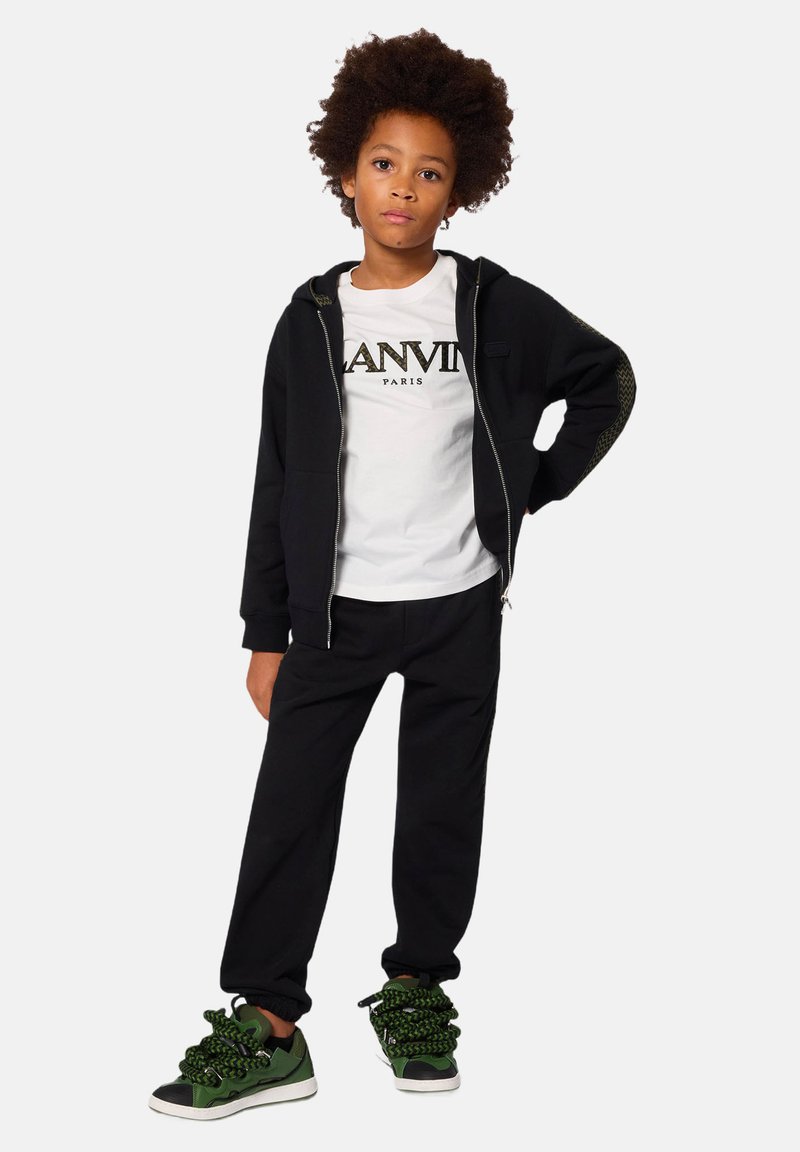 Felpa nera con zip e dettagli in maglia, camicia bianca con "LANVIN PARIS", pantaloni jogger neri e sneakers verdi con lacci spessi.
