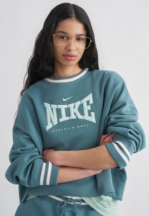 Młoda kobieta z długimi czarnymi włosami, nosząca okulary i turkusową bluzę Nike, stoi z założonymi ramionami na jednolitym tle.