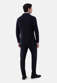 Mann, der mit dem Rücken zur Kamera steht, trägt ein strukturiertes dunkles Blazer, passende Hosen und schwarze Schuhe vor einem einfarbigen hellen Hintergrund.