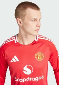 Maillot rouge de Manchester United, avec un col rond, des rayures blanches sur les manches et les logos de l'équipe et de Snapdragon à l'avant.