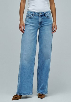 Femme portant un jean taille haute bleu clair à jambes larges, associé à des talons pointus marron et un top blanc sans manches.