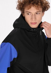 Sort og blå vindjakke med hætte, der har en glat tekstur, halv-zip design og snørrelukning. Bemærkelsesværdige detaljer inkluderer en lynlåstræk.