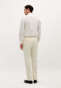 Weißes Langarmshirt mit Standardkragen, kombiniert mit hellfarbigen Hosen. Texturiertes Material, Knopfdetails und schwarze Schuhe.