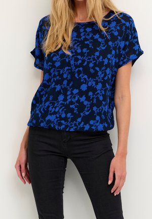 Femme portant un haut noir à manches courtes avec un motif floral bleu vif et un pantalon noir ajusté, debout devant un fond blanc uni.