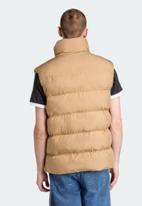 Béžová prošívaná vesta s vysokým límcem, svislým prošitím a vycpávaným designem. Nošená přes tmavé tričko s krátkým rukávem. Hladká textura látky.