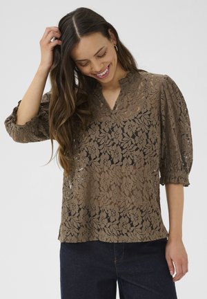 Blouse en dentelle florale marron avec un col en V, des manches bouffantes et une texture semi-transparente, associée à un pantalon en denim sombre.