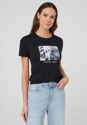 T-shirt in cotone nero con una grafica bianca di un cane e un testo sottostante che recita: "È di nuovo lunedì...?" Modello che indossa jeans azzurri.