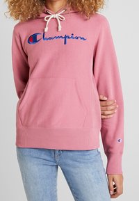 Haut à capuche rose avec cordon de serrage, poche kangourou à l'avant et logo "Champion" brodé en bleu. Tissu doux avec des poignets et un ourlet côtelés.