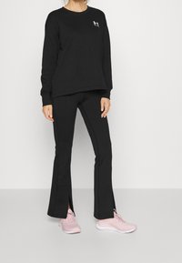 Svart sweatshirt och utsvängda byxor med sidoklyftor, tillsammans med ljusrosa träningsskor. Materialet verkar mjukt med en avslappnad passform.