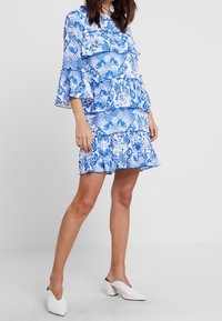 Vestido azul con estampado, con volantes en capas, mangas largas y una falda acampanada. Terminado con una tela ligera y zapatos de tacón cuadrado blancos.