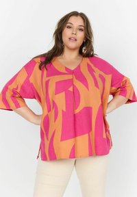 V-halsblouse in oranje en roze stof met abstracte patronen. Losse pasvorm met korte mouwen en zijsplitten. Textuur, lichtgewicht materiaal.