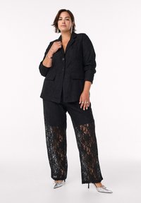 Blazer in pizzo nero con bottoni e tasche, abbinato a pantaloni neri con pannelli in pizzo. Tacchi a punta argentati completano l'outfit.