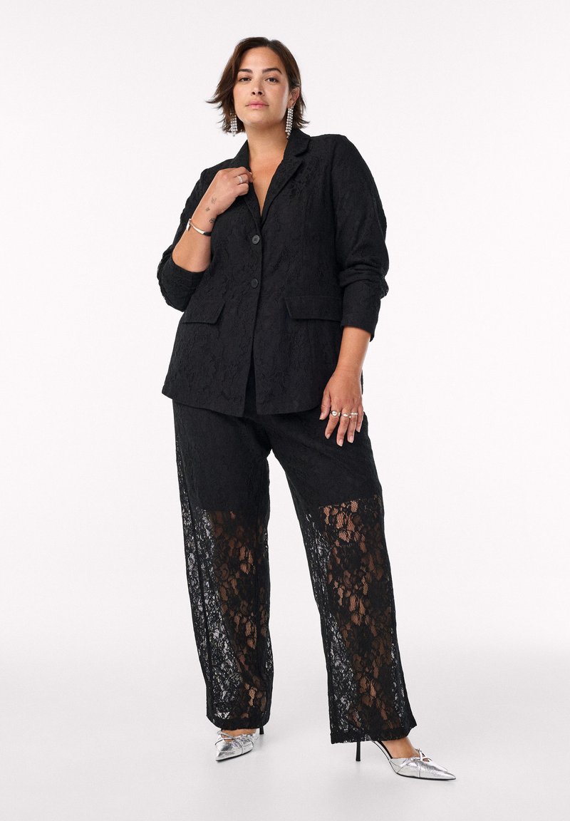 Blazer in pizzo nero con bottoni e tasche, abbinato a pantaloni neri con pannelli in pizzo. Tacchi a punta argentati completano l'outfit.