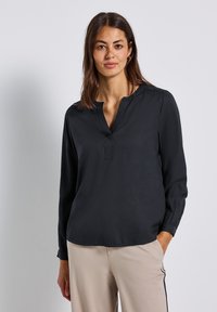 Donna con capelli castani che indossa orecchini a cerchio dorati, una blusa nera a maniche lunghe con scollo a V e pantaloni beige con strisce laterali nere, in piedi contro un muro bianco.