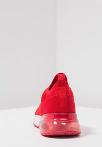 Červená sportovní bota s pleteným svrškem, texturovaným povrchem a průhlednou vzduchovou tlumicí podrážkou. Obsahuje logo na patě a štíhlý, aerodynamický design.