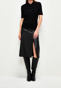 Polo noir en maille côtelée avec manches courtes et fermeture à trois boutons, associé à une jupe midi noire en similicuir avec une fente latérale. Bottes noires montantes jusqu'aux genoux.