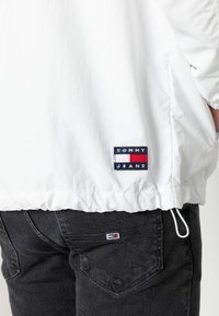 Biała lekka kurtka z elastycznym ściągaczem u dołu, ozdobiona czarno-czerwono-niebieską naszywką z logo "Tommy Jeans". Noszona z ciemnoszarymi jeansami.