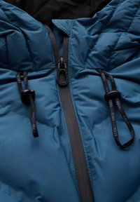 Veste rembourrée turquoise avec une fermeture éclair et des accents noirs, cordons de serrage ajustables et une texture lisse et mate.
