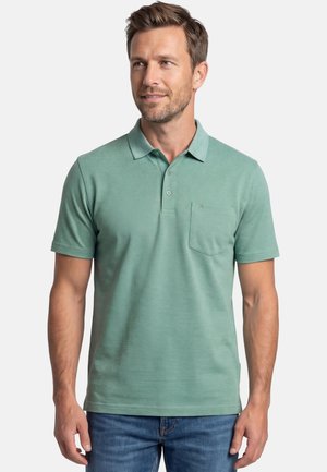 Homme portant un polo vert clair à manches courtes avec une poche poitrine, debout devant un fond blanc uni.