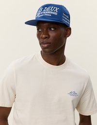 Niebieska czapka z białym napisem, płaski daszek i logo. Jasnobeżowa koszulka z krótkim rękawem z małym niebieskim logo na klatce piersiowej. Gładka tkanina, luźny krój.