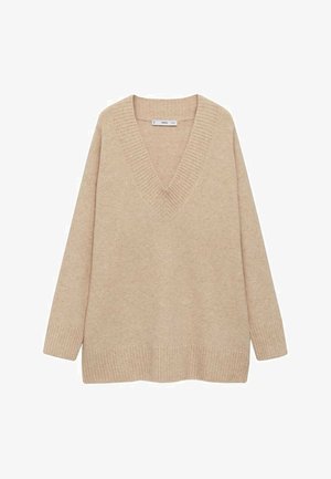 Pull beige à col en V avec poignets côtelés, rentré dans un pantalon noir plissé taille haute, maintenu par une ceinture marron foncé avec une boucle dorée.