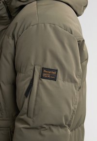 Doudoune vert olive avec une surface texturée, dotée d'une poche zippée et d'un patch de marque noir sur la manche.