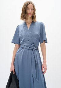 Blaue Wickelkleid mit kurzen Ärmeln, strukturiertem Stoff, tailliert mit einem Knoten-Detail und fließender Silhouette. Das Model hält eine schwarze Jacke.
